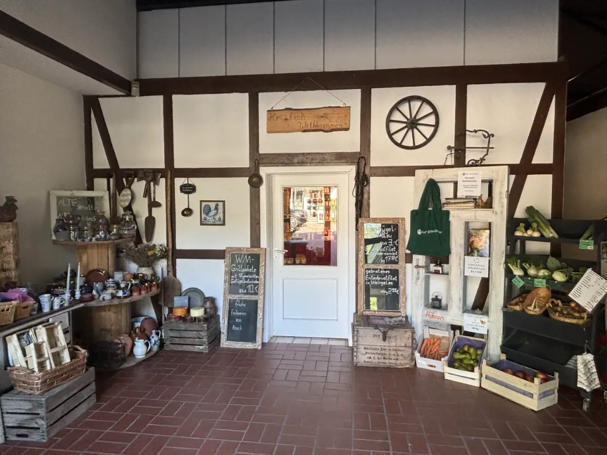 Der Eingangsbereich des Hofladens auf Hof Bimöhlen ist im rustikalen Landhausstil gestaltet. Saisonale Blumen, historische Hofutensilien und dekorative Tafeln schaffen eine einladende Atmosphäre. Durch die Tür ist der Verkaufsraum mit regionalen Produkten sichtbar.