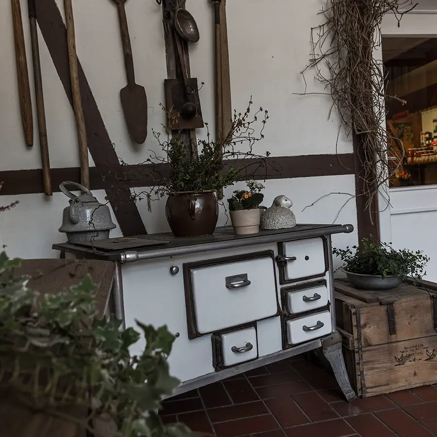 Das Bild zeigt einen alten weißen Küchenherd mit Emailleoberfläche, dekoriert mit Pflanzen und traditionellen Küchenutensilien. An der Fachwerkwand hängen historische Werkzeuge und Haushaltsgegenstände. Die rustikale Einrichtung vermittelt die gemütliche Atmosphäre des traditionellen Bauernhofs Bimöhlen.