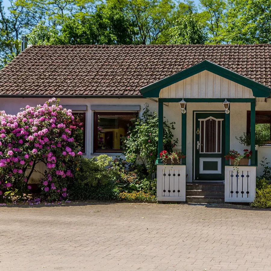 Das Foto zeigt ein gepflegtes Bauernhaus mit überdachtem Eingangsbereich auf Hof Bimöhlen. Vor dem Gebäude blüht ein großer Rhododendronstrauch in kräftigem Rosa. Die Hofanlage wirkt einladend und ist von Bäumen und weiteren Pflanzen umgeben.