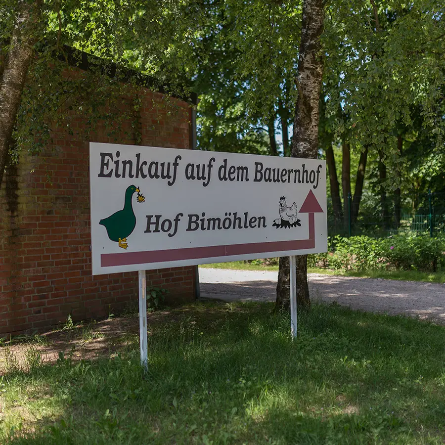 Ein großes Hinweisschild mit der Aufschrift „Einkauf auf dem Bauernhof – Hof Bimöhlen“ steht am Straßenrand zwischen Bäumen und zeigt Besuchern den Weg zum Hofladen. Die ländliche Umgebung mit Grünflächen und Baumbestand unterstreicht den Charakter eines regionalen Bauernhofs.