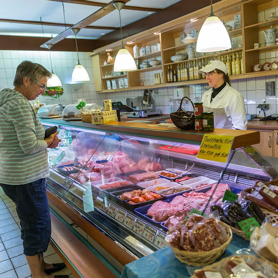 Das Foto zeigt den Verkaufsraum des Hofladens vom Hof Bimöhlen. Eine Kundin steht vor einer gut gefüllten Frischetheke mit Fleisch- und Wurstwaren, während eine Mitarbeiterin hinter dem Tresen berät. Im Hintergrund befinden sich Regale mit regionalen Produkten und hofeigenen Spezialitäten.