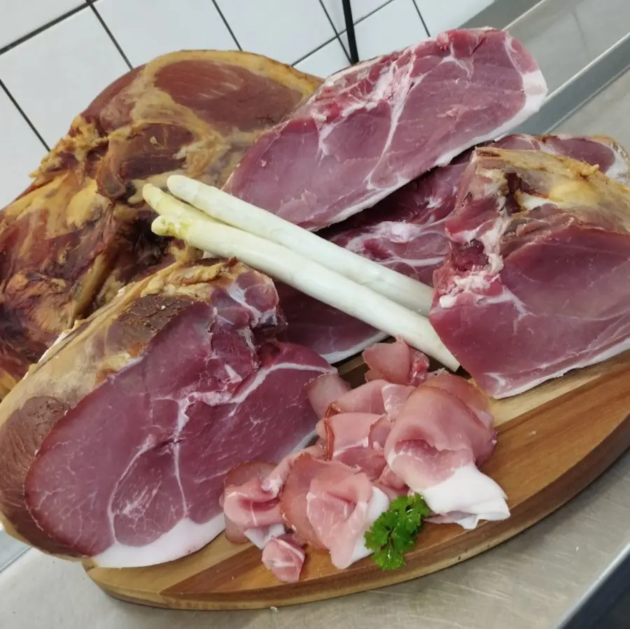 Mehrere große Stücke geräucherter Schinken liegen auf einem Holzbrett. Im Vordergrund befinden sich dünn aufgeschnittene Schinkenscheiben, dekoriert mit frischer Petersilie. Die Aufnahme zeigt die handwerkliche Verarbeitung und hochwertige Qualität der Schinkenspezialitäten vom Hof Bimöhlen.
