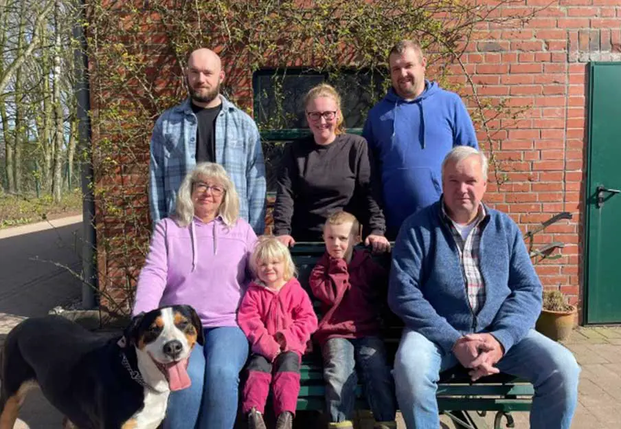 Das Foto zeigt mehrere Generationen der Familie von Hof Bimöhlen vor einem Backsteingebäude auf dem Hofgelände. Erwachsene, Kinder und ein Hund sind gemeinsam auf einer Bank und davor gruppiert. Die Aufnahme vermittelt den familiären Charakter des landwirtschaftlichen Familienbetriebs.