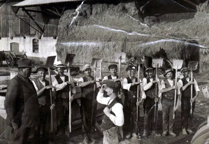 Das Schwarz-Weiß-Foto zeigt eine Gruppe von Landarbeitern auf einem Bauernhof. Die Männer stehen mit traditionellen Arbeitsgeräten vor einem großen Heu- oder Strohlager und dokumentieren die landwirtschaftliche Arbeit vergangener Generationen.