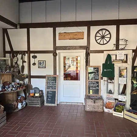 Der Eingangsbereich des Hofladens auf Hof Bimöhlen ist im rustikalen Landhausstil gestaltet. Saisonale Blumen, historische Hofutensilien und dekorative Tafeln schaffen eine einladende Atmosphäre. Durch die Tür ist der Verkaufsraum mit regionalen Produkten sichtbar.